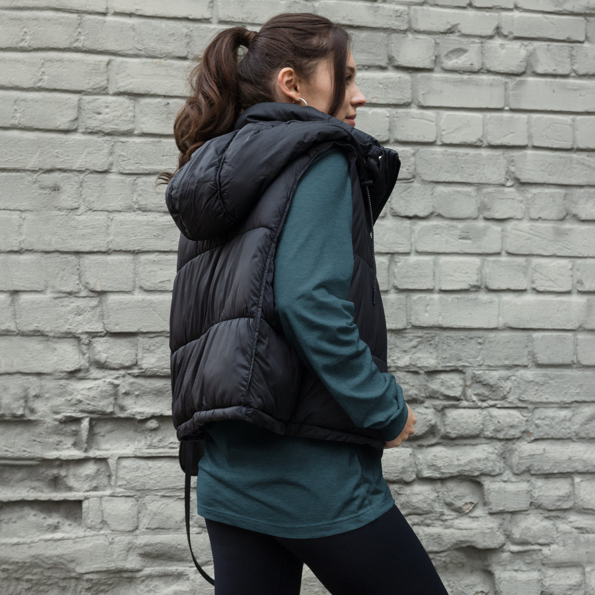 Model wearing a heather deep teal TMiTH long sleeve tee under a black padded vest, back and side view; the back design is covered. The Man in The Hat 907 artist merch expressing PTSD and complex trauma expression.