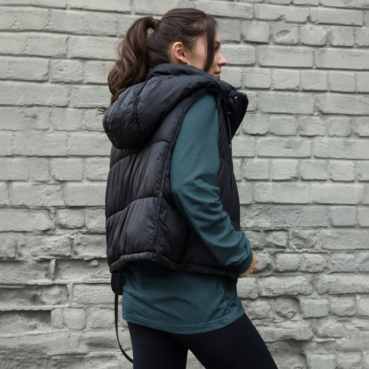 Model wearing a heather deep teal TMiTH long sleeve tee under a black padded vest, back and side view; the back design is covered. The Man in The Hat 907 artist merch expressing PTSD and complex trauma expression.